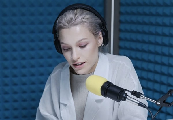 Close up view of woman with headphones looking down, speaking into mic in a blue soundproof recording room