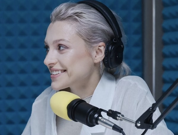tile-View of two women facing each other, both wearing headphones with their own mic in a blue soundproof room, chatting and smiling