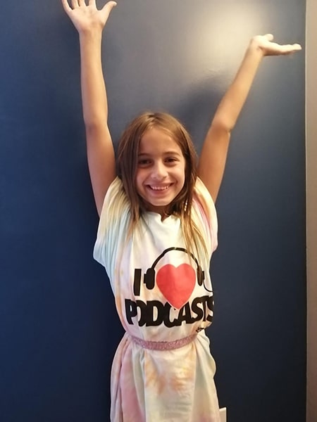 a 8-year-old blonde smiling girl in a shirt with I love podcasts and having her hands up-min