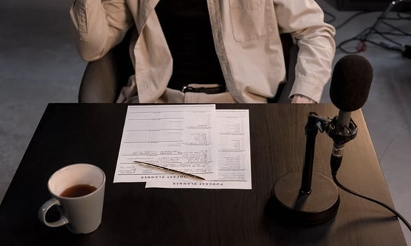 Angled down view of person sitting at desk with podcast notes, a mic, and cup of coffee in recording studio