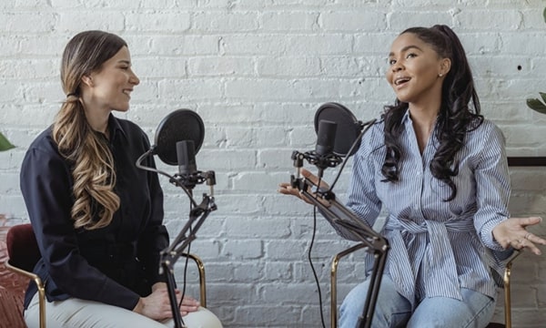 Two young women against a white brick background sitting separately, chatting and smiling into microphones for a podcast interview