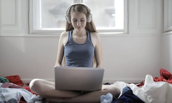 View of smiling woman sitting cross-legged in room of clothing on floor wearing headphones and diligently working on laptop