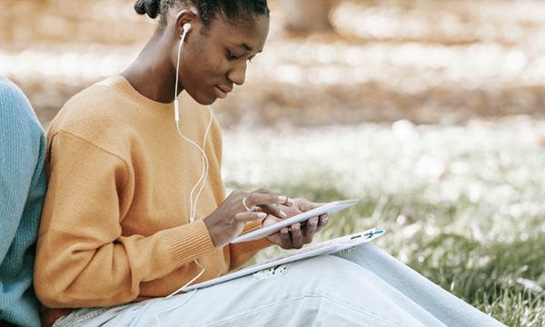 Woman sitting outdoors on grass, leaning against another person’s back, wearing headphones, and working on a tablet